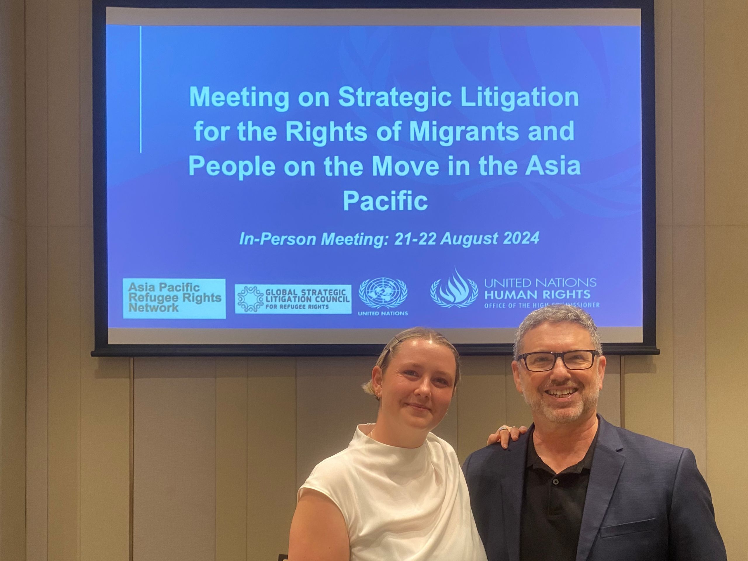 Two people standing in front of a powerpoint slide that reads "meeting on strategic litigation for the rights of migrants and people on the move in the asia pacific